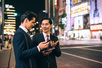 Japanese businessmen explaining something to each other and holding a tablet outside in the city.
